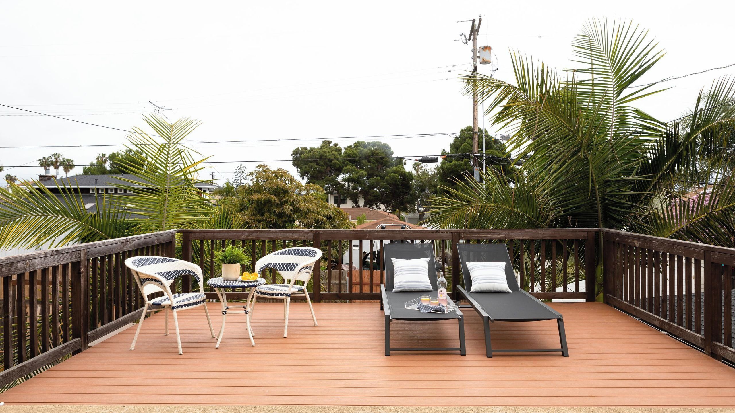 Une terrasse en bois avec deux chaises longues et une table, entourée de palmiers et offrant une vue sur les maisons.