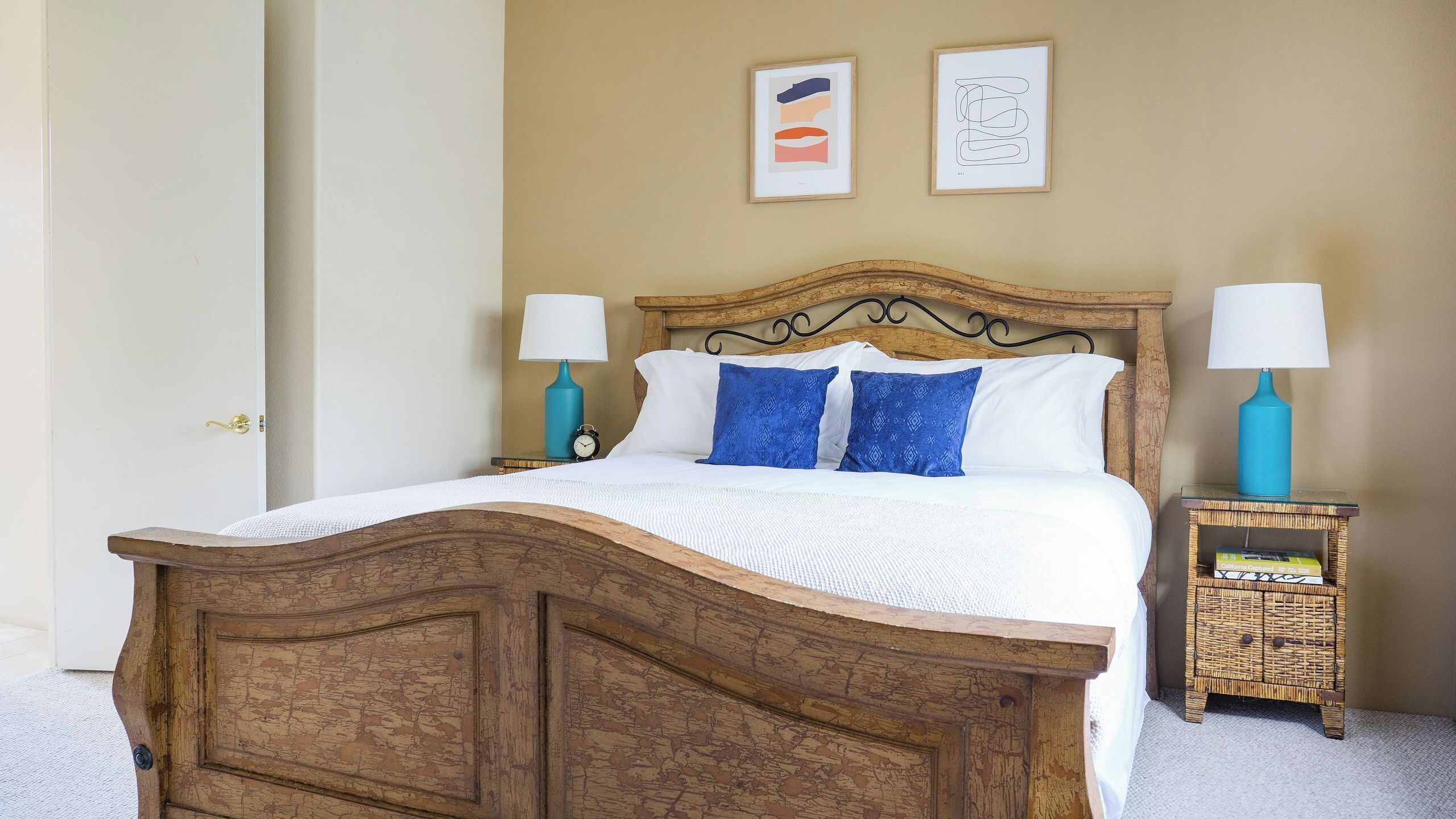 A wooden bed with white bedding, two blue pillows, a nightstand with a lamp, and a wicker side table with a lamp.