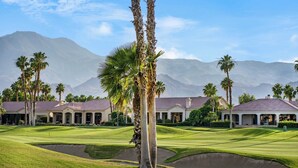 Een golfbaan met palmbomen, een clubhuis en bergen op de achtergrond.