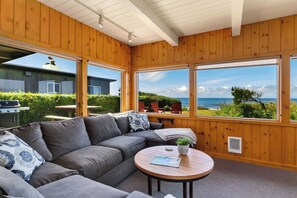 A cozy room with a wooden interior, a gray sofa, a round table, and large windows offering a view of the sea and greenery. - The Pilot House by Avantstay Stunning Home Right On Arch Cape Beach! (Arch Cape)