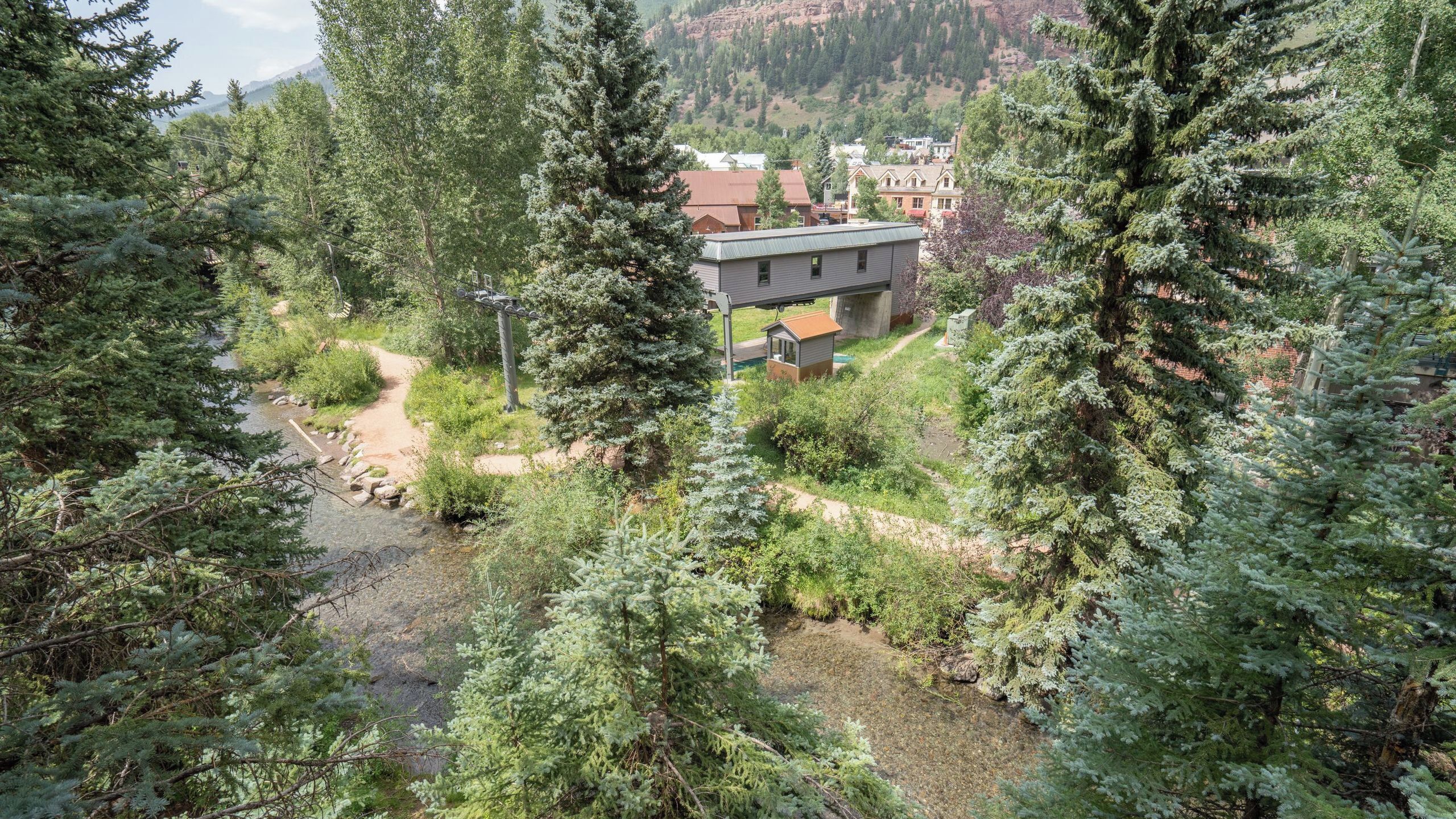 A modern building with a flat roof, surrounded by dense forest and a small stream.