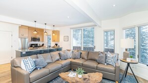 A modern living room with a sectional sofa, a wooden coffee table, and a kitchen area with wooden cabinets and pendant lighting. - Manitou Riverhouse 115 by Avantstay Spacious Condo in the Centre of Telluride (Telluride)