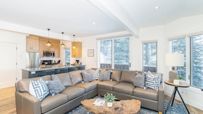 A modern living room with a sectional sofa, a wooden coffee table, and a kitchen area with wooden cabinets and pendant lighting. - Manitou Riverhouse 115 by Avantstay Spacious Condo in the Centre of Telluride (Telluride)