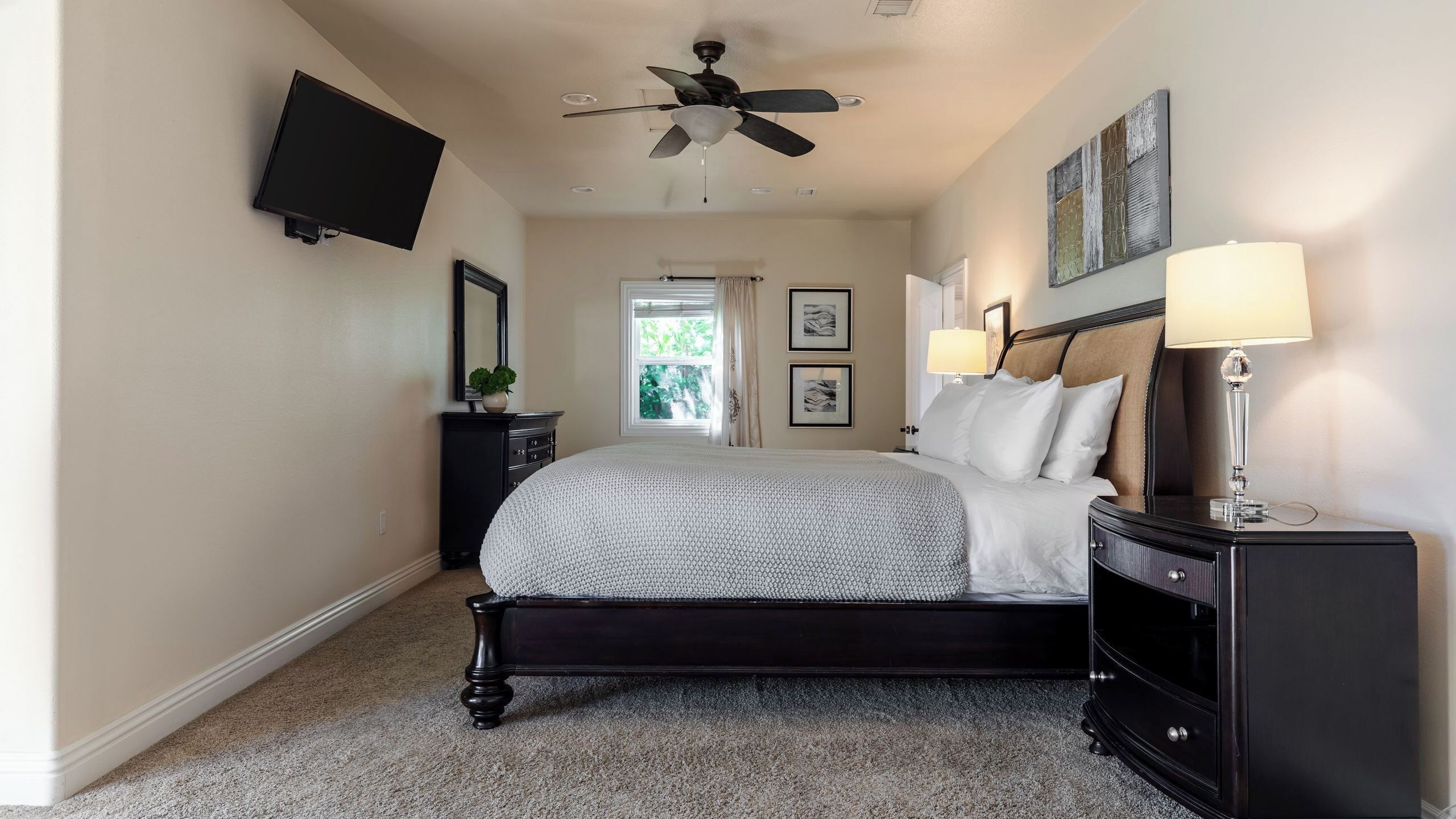 A bedroom with a bed, a ceiling fan, a TV, a nightstand, a lamp, and artwork on the wall.