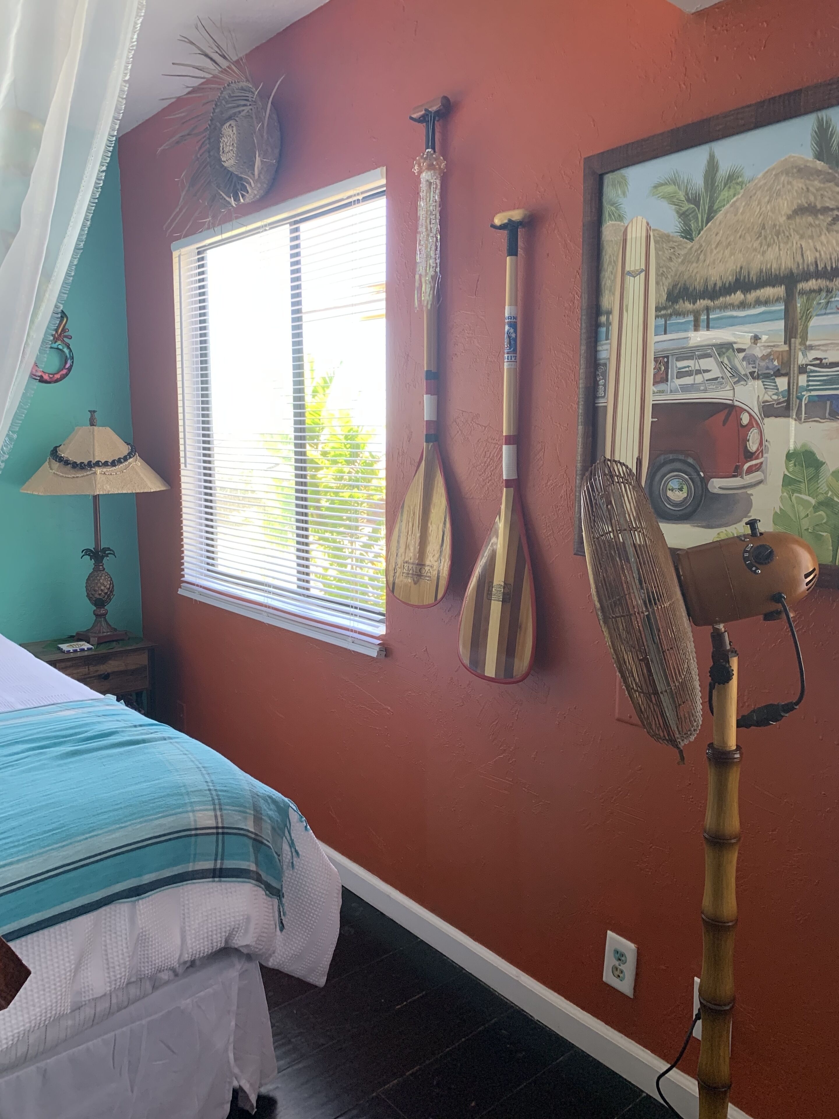 Una habitación con cama, ventana con persianas, lámpara y elementos en la pared, como una pala, un ventilador y un cuadro.
