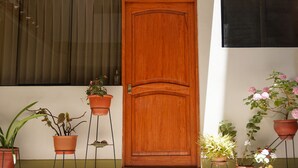 Eine Holztür mit Messingklinke, beides von Blumentöpfen und einem Fenster mit vertikalen Rollläden eingerahmt.