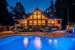 A two-story wooden cabin with a swimming pool, surrounded by a wooden deck and a fenced yard.