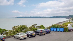 Un espace de stationnement avec des voitures et des poubelles de recyclage bleues, offrant une vue sur une étendue d’eau et une côte lointaine.