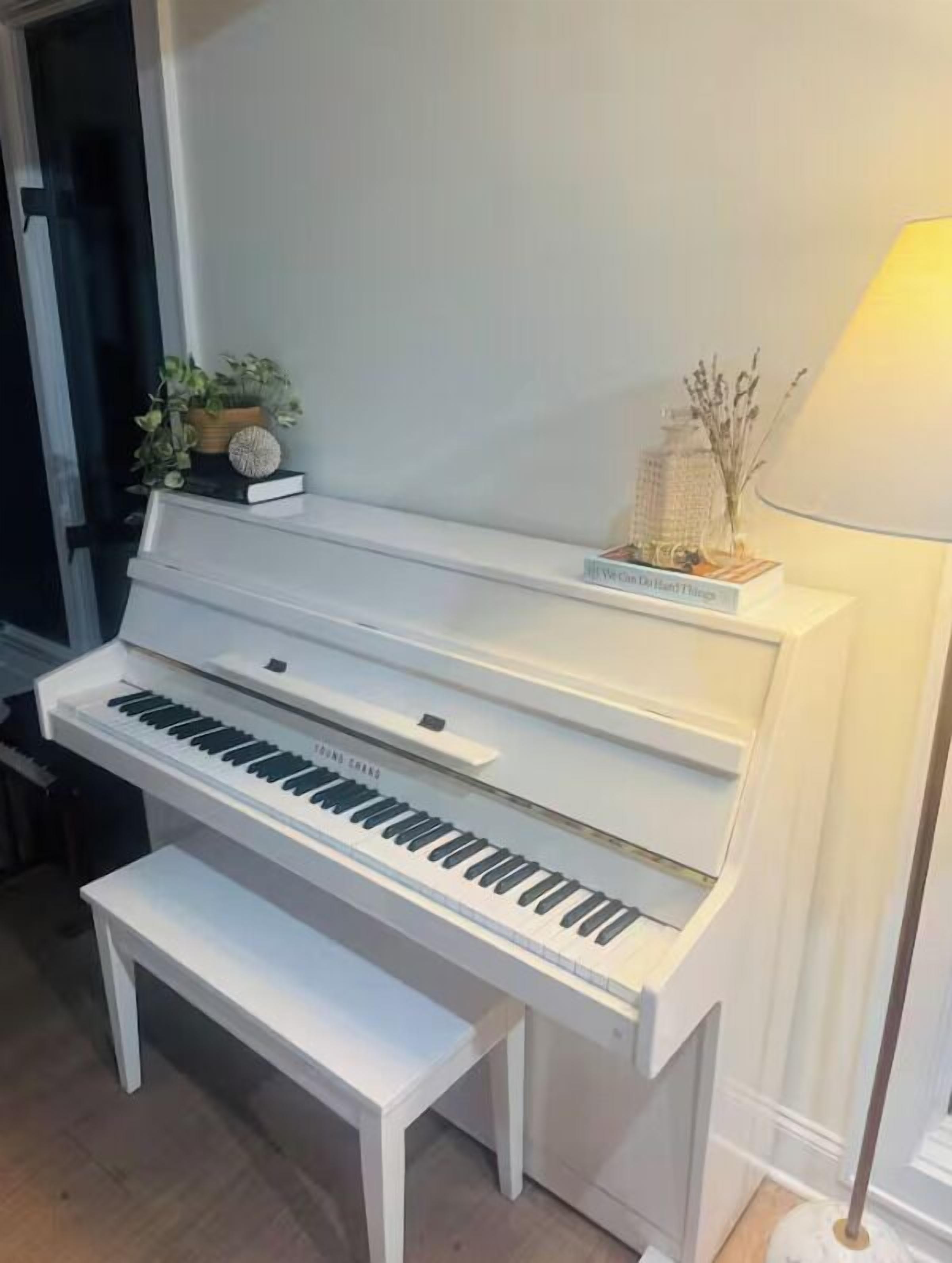 Ein weißes, stehendes Klavier mit einer Bank davor, einer Lampe und dekorativen Gegenständen auf einem Regal.