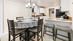 A dining area with a round table set for four, surrounded by dark wooden chairs. A chandelier hangs above the table, and a kitchen area is visible in the background with white cabinets and a countertop. - 2Min>RedhawkGolfCourse★LongStays★300mbps★1300ft²★ (Nampa)