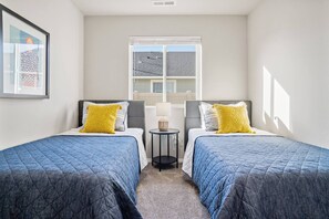 A bedroom with two beds, each with a blue quilted bedspread and a yellow pillow. There is a small round table with a lamp between the beds.