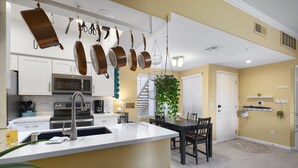 A modern kitchen with white cabinets, stainless steel appliances, and hanging pots. There is a dining area with a table and chairs, and a large plant in the center. - Cozy Desert Getaway*Inviting Pool*Smart TV*Gym (Fountain Hills)