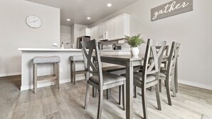 A modern kitchen with white cabinets, a wooden dining table with four chairs, a small island with stools, and a 'gather' sign on the wall. - *FirePit*King*Parking*1600ft²*Smart TV*W/D (Meridian)
