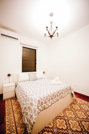 Une chambre à coucher comprenant un lit, une table de chevet, une commode, une fenêtre avec des stores et un lustre.