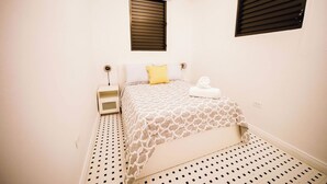 Une petite chambre propre, avec un lit, une table de chevet, une fenêtre avec des stores et un appareil de climatisation.