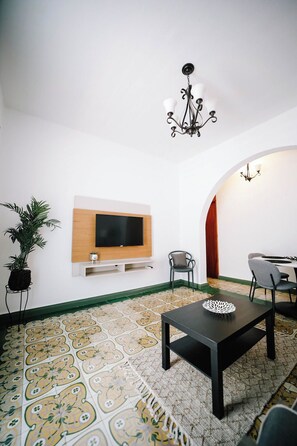 A living room with a flat-screen TV mounted on a wooden panel, a black coffee table with a decorative item, a potted plant, and a patterned rug. - Colonial Apartment, Generator, Walk to San Juan (San Juan)