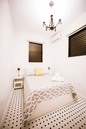 A bedroom with a bed, a nightstand, a wall-mounted air conditioner, and a window with blinds.