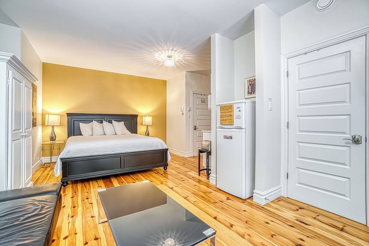 A bedroom with a bed, bedside tables, a desk, a refrigerator, and a door.