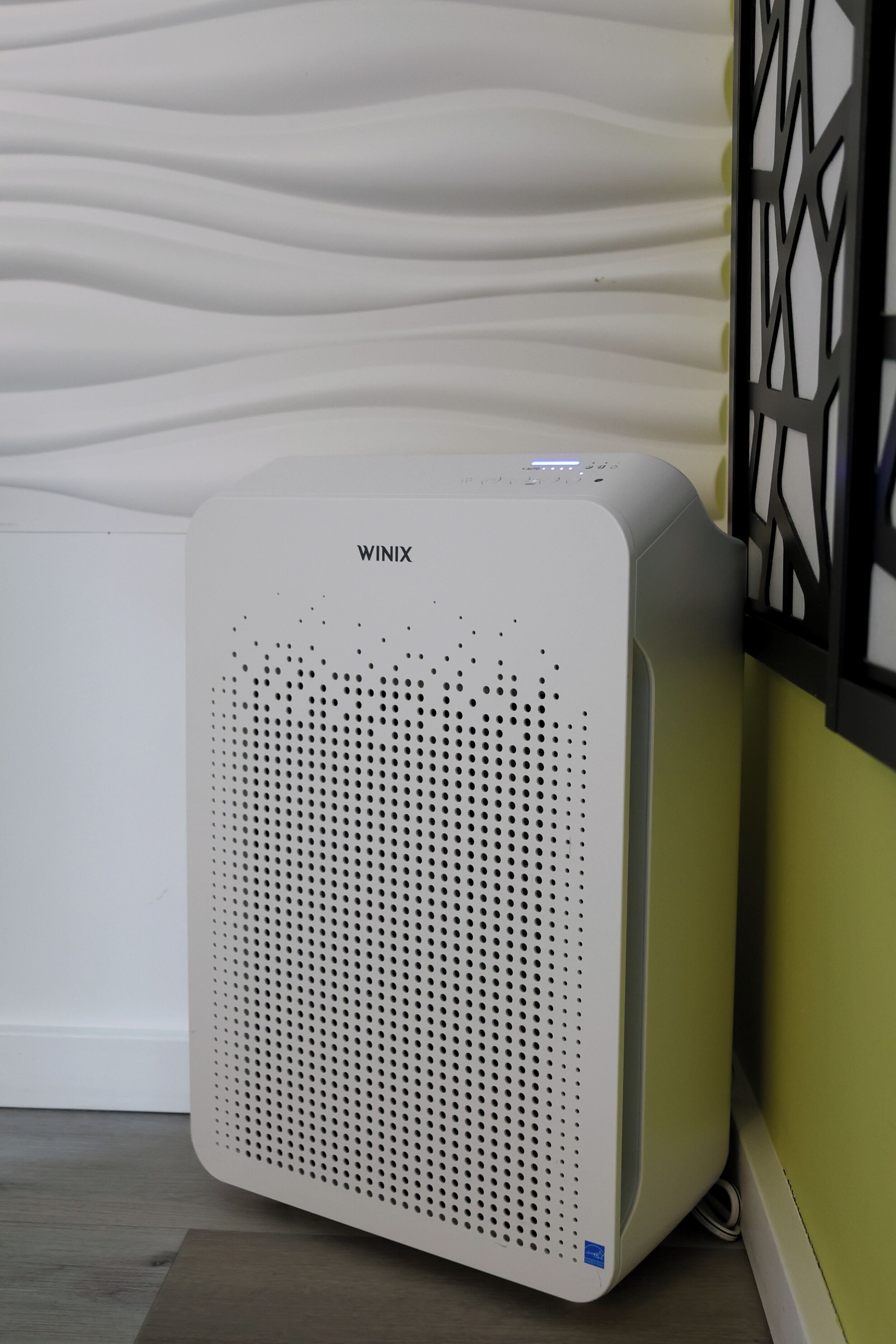 A white air purifier with the brand name WINIX, placed on a wooden floor against a wall with a geometric pattern.