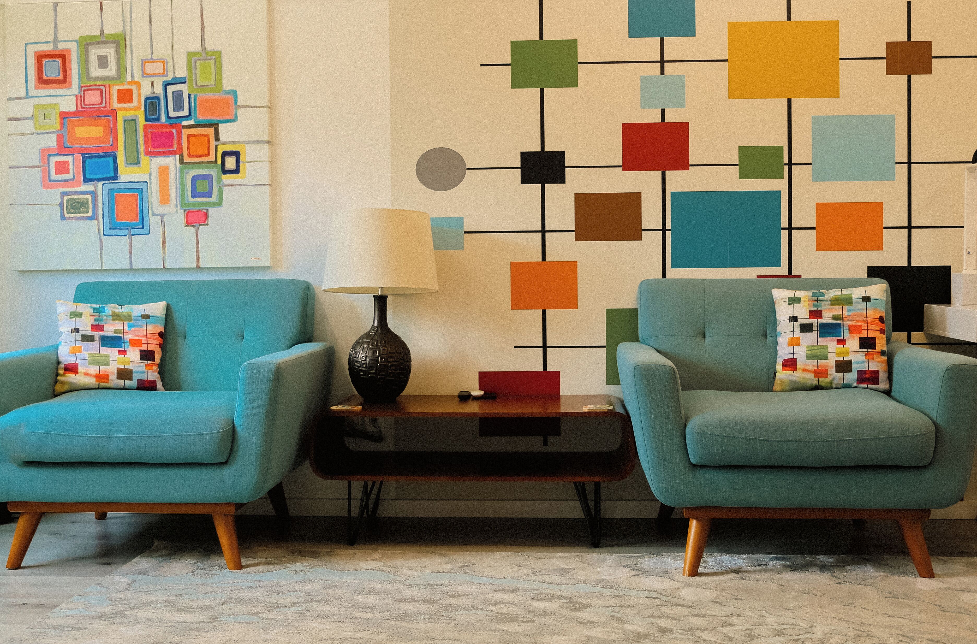 Dos sillones azul turquesa con cojines estampados, una mesita con una lámpara y una pared decorada con arte geométrico.