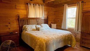 Un dormitorio de cabaña de madera con cama, mesita de noche, ventilador y ventana con cortinas.