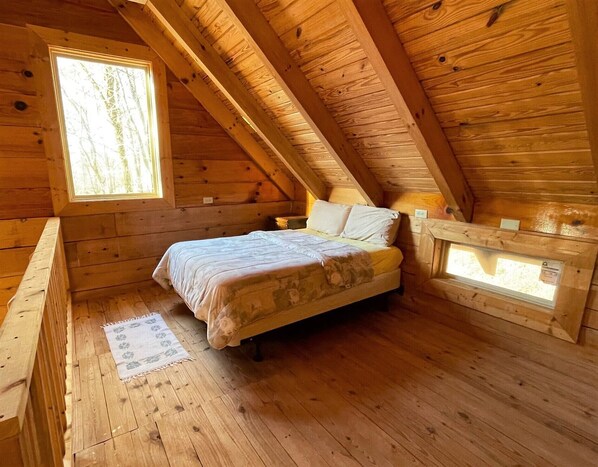 Habitació d'una cabana de fusta amb un llit, una finestra i una taula petita.