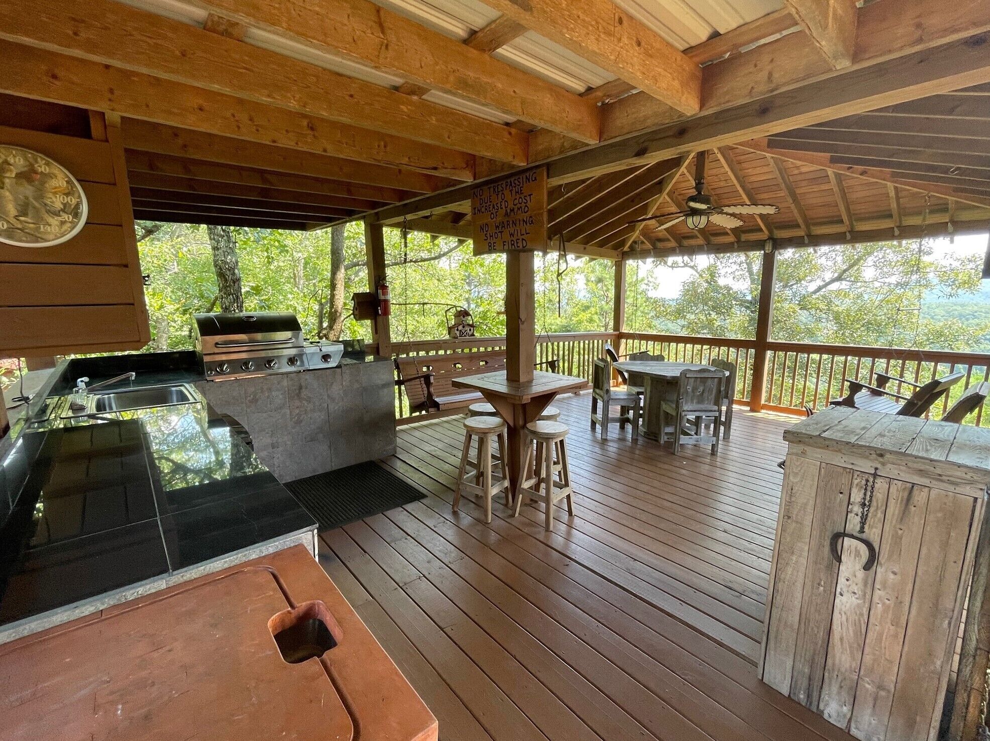 An outdoor deck with a barbecue area, wooden tables and chairs, and a view of the forest.
