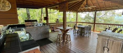 An outdoor deck with a barbecue area, wooden tables and chairs, and a view of the forest.