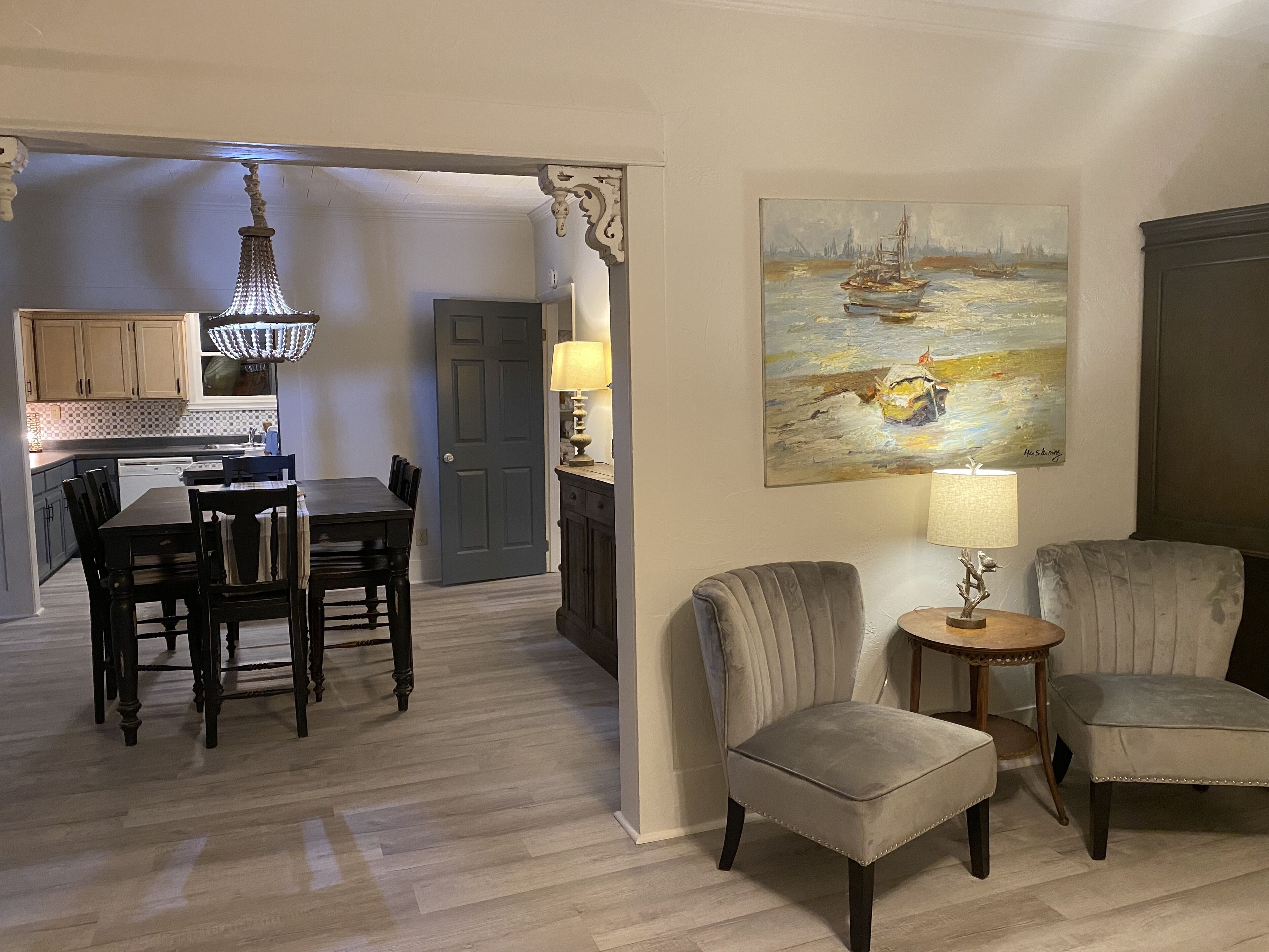 A dining area with a chandelier, a painting of a boat on water, and a door leading to a kitchen.