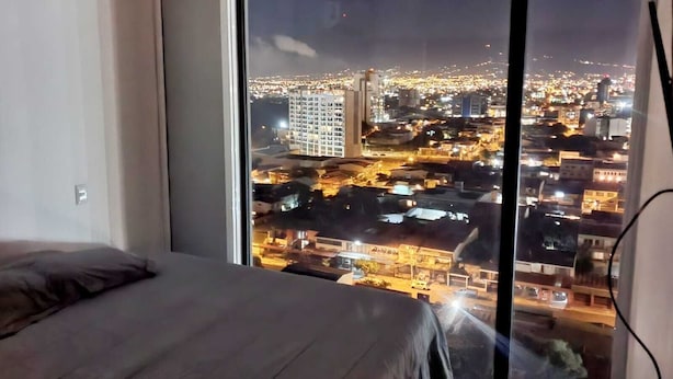 A bedroom with a large window overlooking a cityscape at night. - Luxurious apartment one block from the National Stadium (San José)
