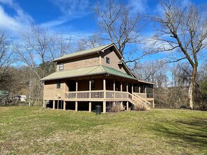 A two-story wooden cabin with a balcony and a steep roof, surrounded by trees and a grassy area.