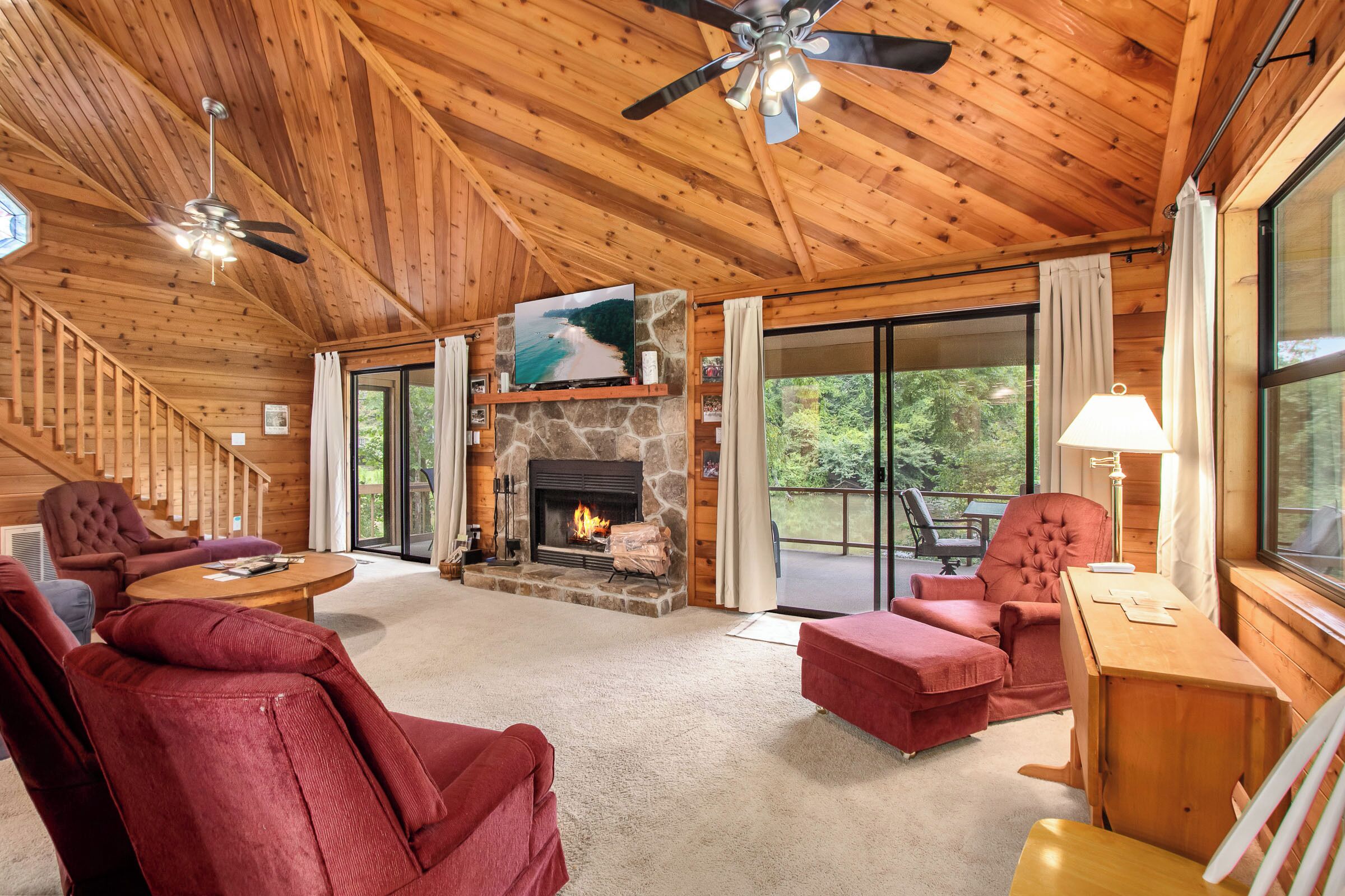 A log cabin interior with a stone fireplace, wooden ceiling, and large windows.