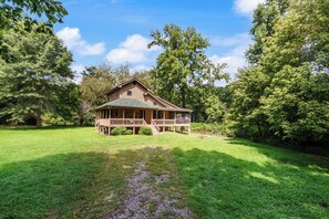 A two-story wooden house with a porch, surrounded by lush greenery and trees.
