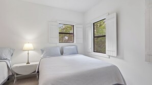 Ein Schlafzimmer mit einem Bett, zwei Nachttischen, einer Lampe und zwei Fenstern mit Rollläden.