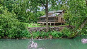 A cabin with a deck, surrounded by dense greenery and a river.