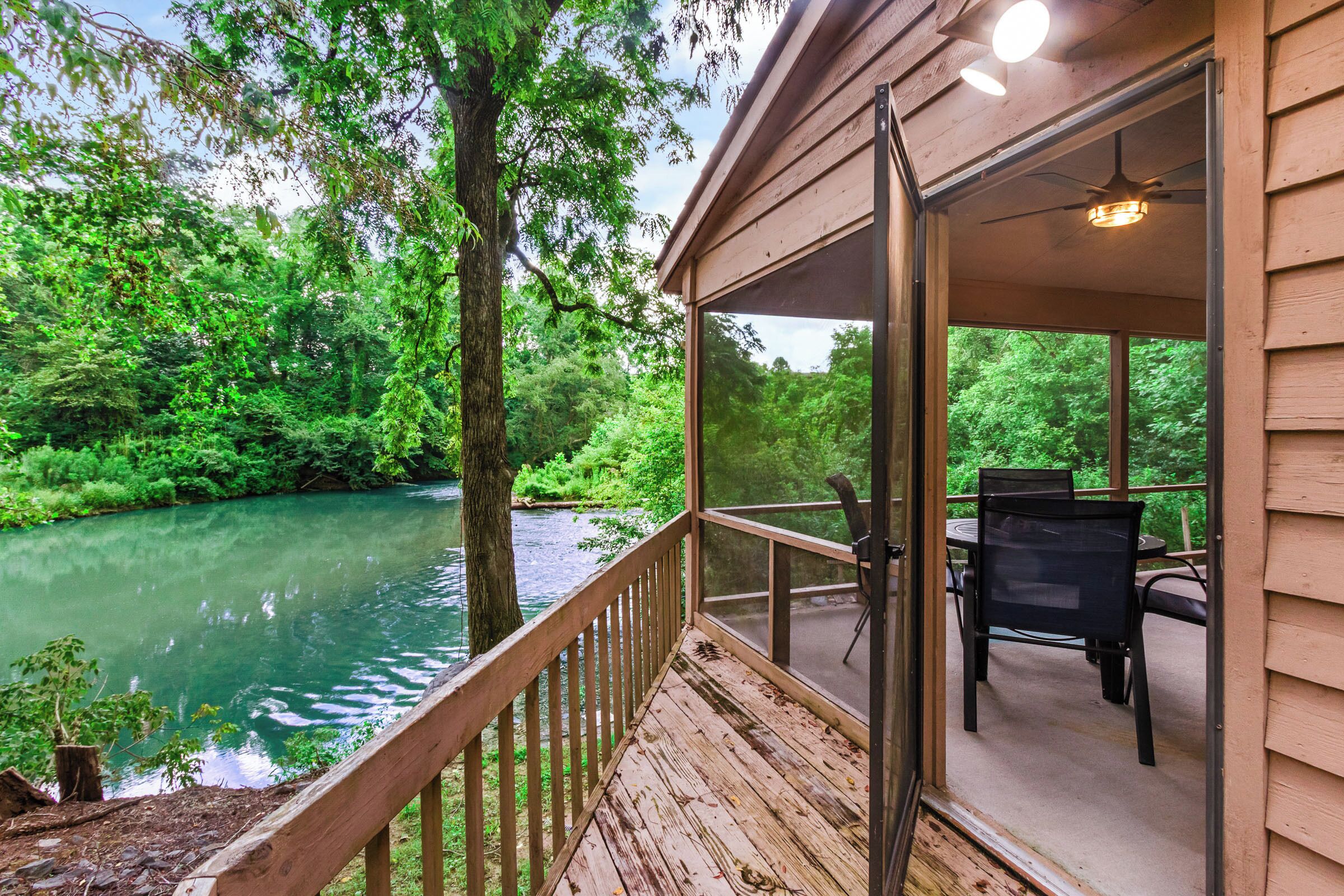 A wooden deck with a glass door leading to an outdoor seating area with a table and chairs, overlooking a river and dense greenery.