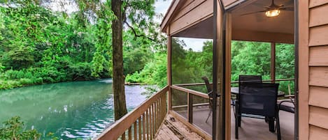 A wooden deck with a glass door leading to an outdoor seating area with a table and chairs, overlooking a river and dense greenery.
