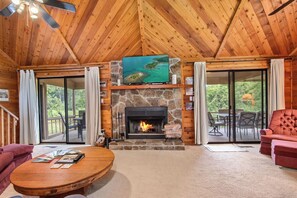 A cozy cabin interior with a stone fireplace, a television mounted above it, and a large window offering a view of the outdoors.