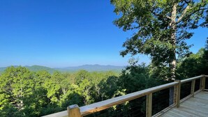 Um deck de madeira com guarda-corpo de metal, com vista para uma paisagem arborizada e montanhas ao longe.