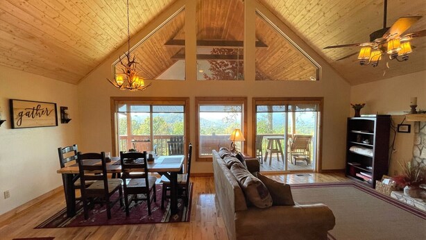 Um amplo loft com teto alto, vigas de madeira, área de jantar com mesa e cadeiras, um sofá e grandes janelas com vista para o exterior.