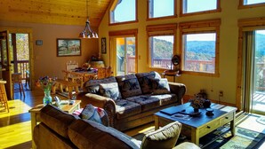 Ein geräumiges Wohnzimmer mit hohen Decken, Holzbalken, einem großen Sofa, einem Esstisch und mehreren Fenstern mit Blick ins Freie.