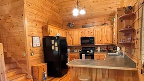 Ein Interieur einer Holzhütte mit einer Küche, die einen schwarzen Kühlschrank, eine Mikrowelle und Holzauszüge beinhaltet. An der Küchenarbeitsplatte stehen zwei Hocker und darunter befindet sich ein kleiner Holzauszug.