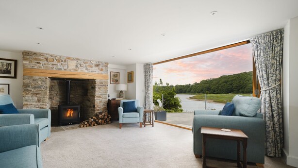 A cozy living room with a stone fireplace, two blue armchairs, a small table, and a view of a river and greenery.