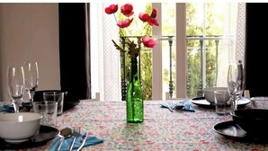 Une table à manger dressée avec des assiettes, des bols, des verres et des couverts, et un vase vert rempli de fleurs placé au centre.