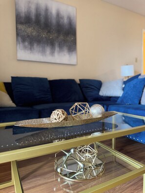 A modern living room with a glass coffee table, a blue sofa, and a painting on the wall.