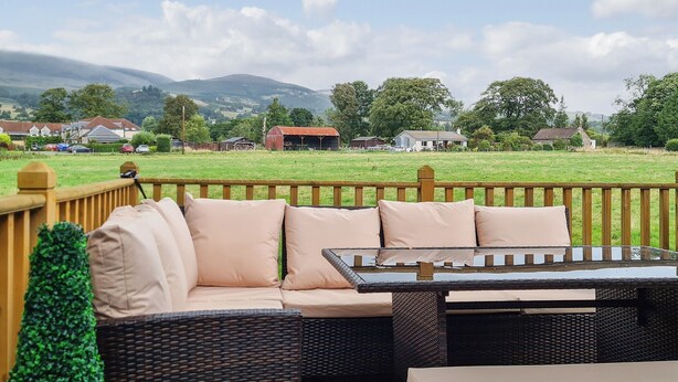 A wicker sofa with cushions and a glass-top table on an outdoor deck with a view of a green field and houses in the distance. - Castle Campbell View (Dollar)