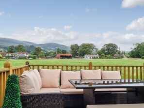 A wicker sofa with cushions and a glass-top table on an outdoor deck with a view of a green field and houses in the distance. - Castle Campbell View (Dollar)