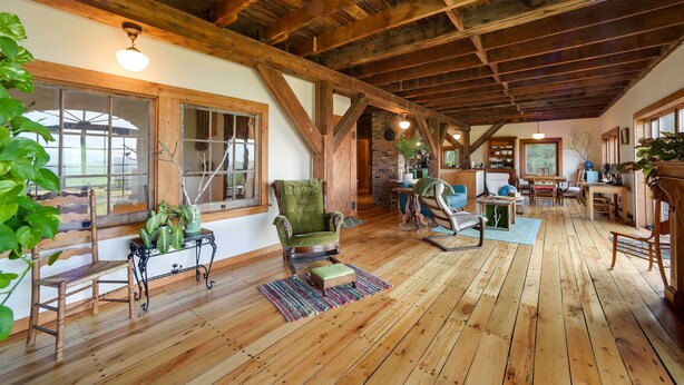 Een ruime kamer met houten vloeren, een hoog plafond met zichtbare balken, een groene fauteuil, een houten schommelstoel en een klein tafeltje met een plant.