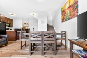 A modern kitchen with wooden cabinets, a dining area with a wooden table and chairs, a flat-screen TV, and a staircase leading to another level.