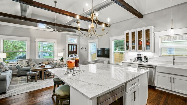 A modern kitchen with a large island, white cabinetry, and stainless steel appliances. There is a dining area with a gray sofa and a chandelier. - Heated Pool, King Beds, EV Charger, Garage & 3BR Sleep 7 · Walk to Beach (Galveston)